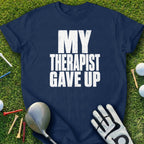 Navy t-shirt with white text reading "MY THERAPIST GAVE UP" on grass with golf equipment.