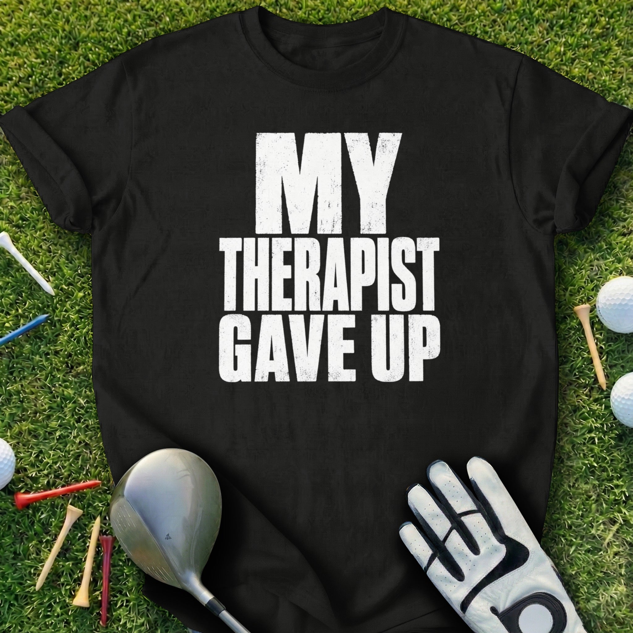 Black t-shirt with white text "MY THERAPIST GAVE UP" on grass with golf clubs.