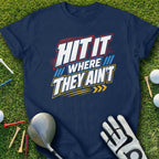 Navy t-shirt with "HIT IT WHERE THEY AIN'T" slogan and golf accessories on grass.