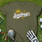 Green t-shirt with "Sandbagger" text and a crown graphic, on grass with golf clubs and balls.