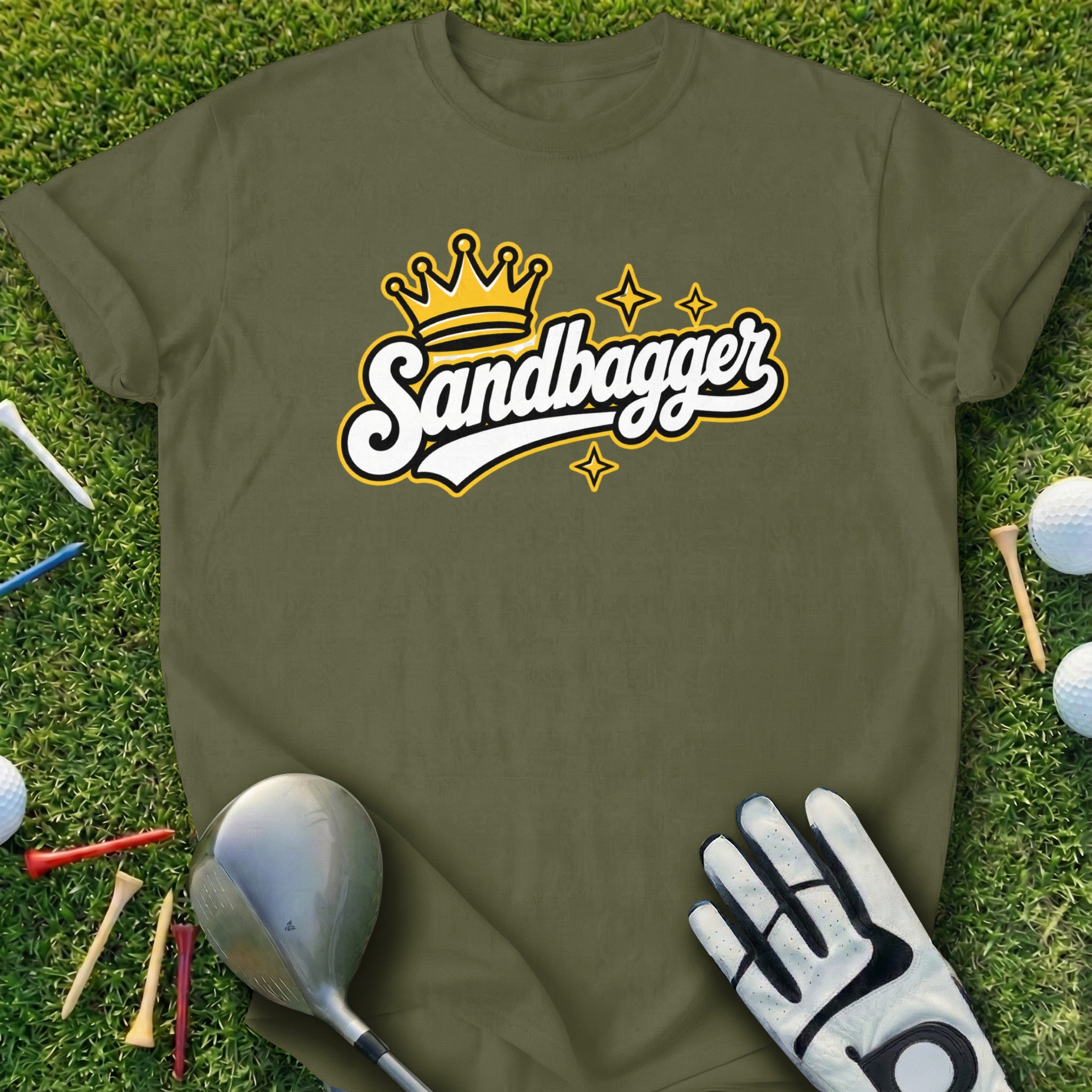 Green t-shirt with "Sandbagger" text and a crown graphic, on grass with golf clubs and balls.