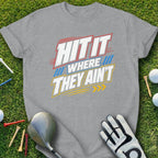 Gray t-shirt with "HIT IT WHERE THEY AIN'T" text, golf glove, tees, and balls on grass.