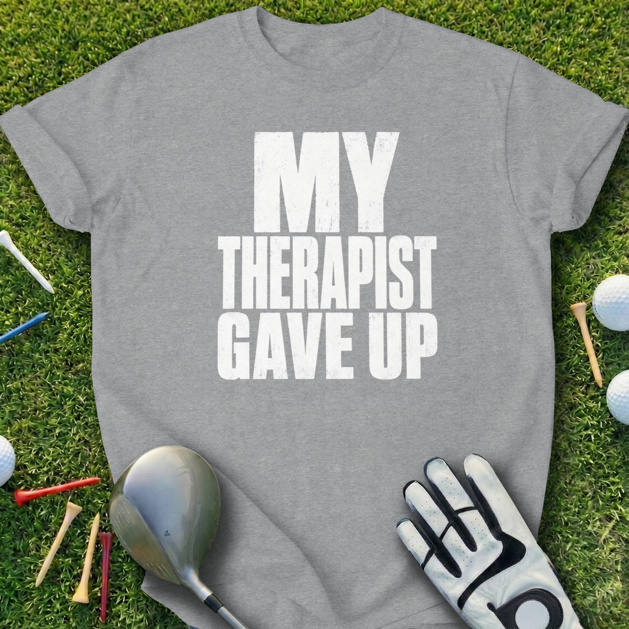 Grey t-shirt with white text "MY THERAPIST GAVE UP" on grass with golf equipment.