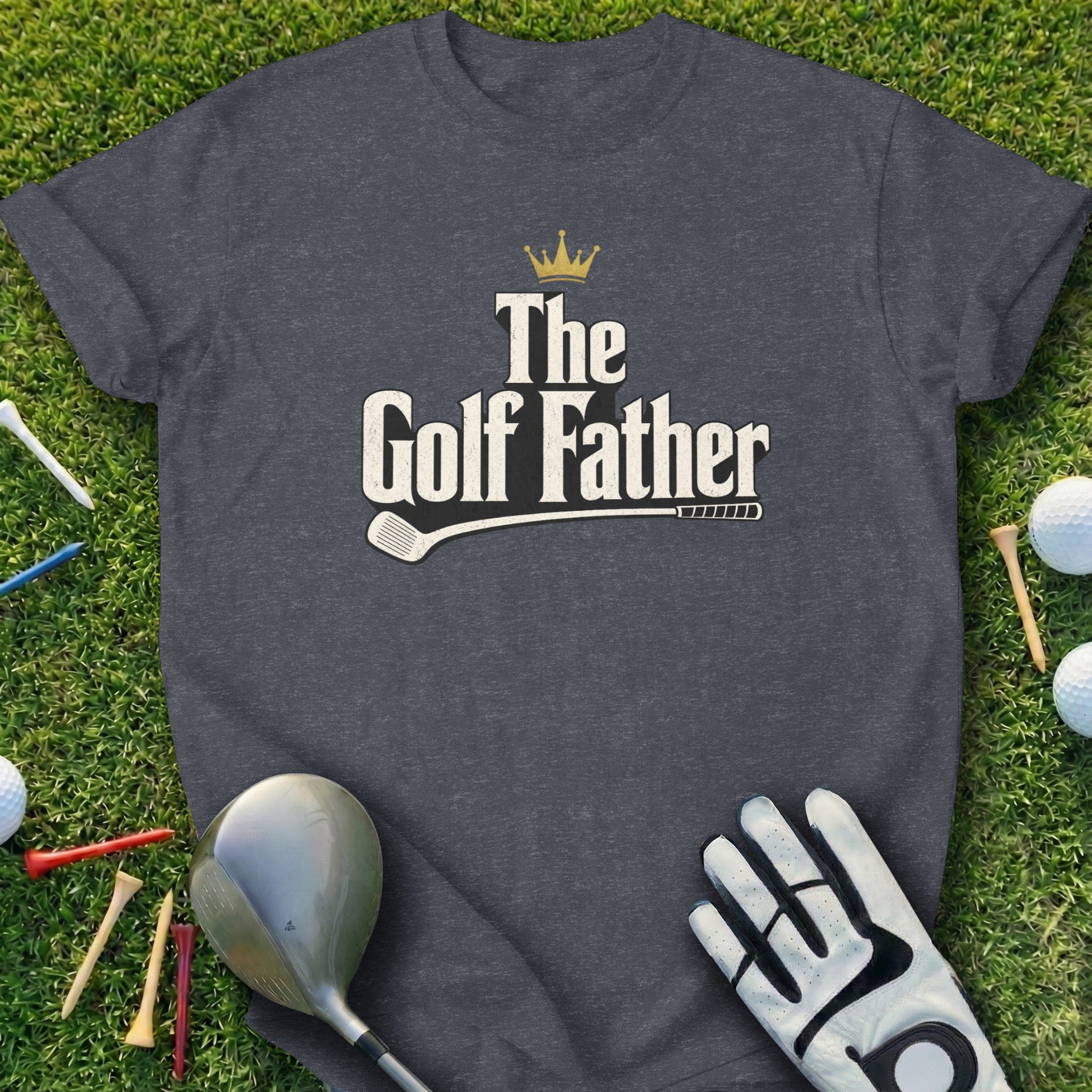 Dark gray t-shirt with "The Golf Father" and crown graphic, on grass with golf items.